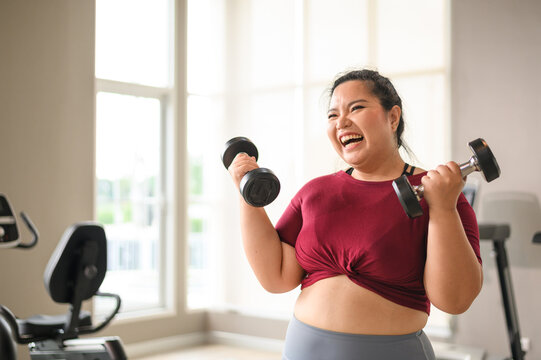 Young, Positive, Smiling, Cheerful overweight woman training with dumbbells in gym. Fat woman in sportswear doing fitness exercise. Weight loss workout, healthy lifestyle concept.