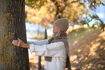 Portrait of a beautiful senior woman hugging tree trunk with closed eyes. Elderly woman in connection with nature.