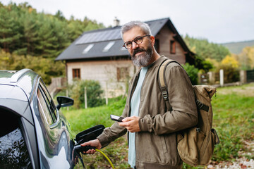 Man charging electric car in front of his house, plugging the charger into the charging port. House...