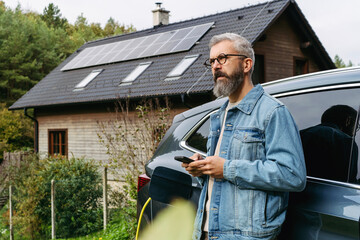 Man charging electric car in front of his house, plugging the charger into the charging port. House...