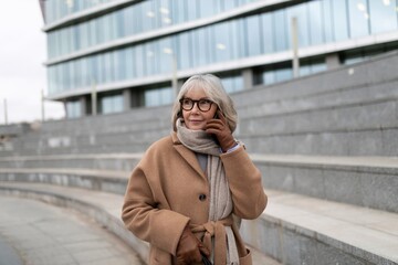 Fototapeta premium Elderly woman wearing glasses and a brown coat talks on the phone while standing outside an urban building during a cloudy day