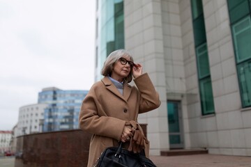 Fototapeta premium An older woman in a brown coat adjusts her glasses while walking near modern buildings on a cloudy day in the city
