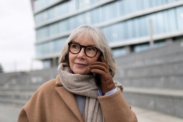Fototapeta premium A senior woman wearing glasses talks on her smartphone while standing outside a modern building on a cool day