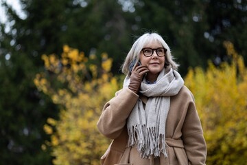 Elderly woman in a beige coat and scarf enjoying a phone call in a park surrounded by autumn leaves during a sunny day