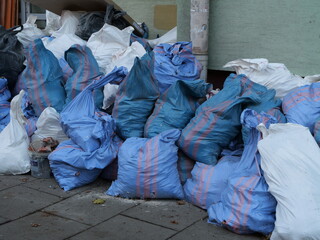 Piles of blue and white garbage bags stacked on a city sidewalk against a building wall, representing urban waste, cleanup, or construction debris removal