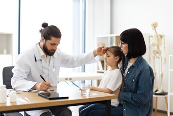 Caucasian male doctor measures temperature by touching forehead of young Caucasian girl in clinic while writing prescription for mother. Girl sits on mother's lap.
