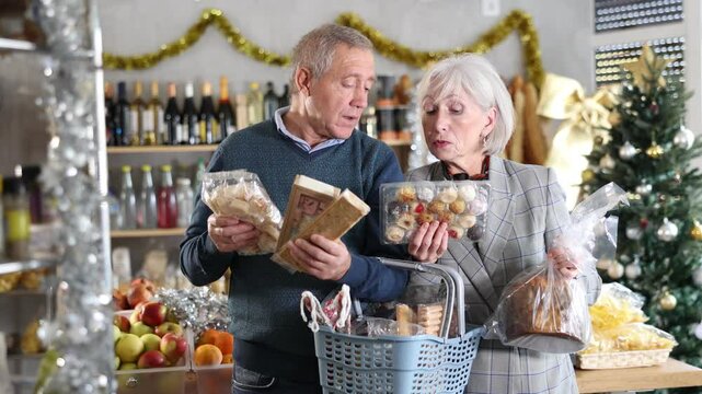 Elderly spouses looking at sweets in store. Market visitors choose consider nougat, panettone, marzipan, cookies, chocolate. Customers choose national European sweet desserts in sales area