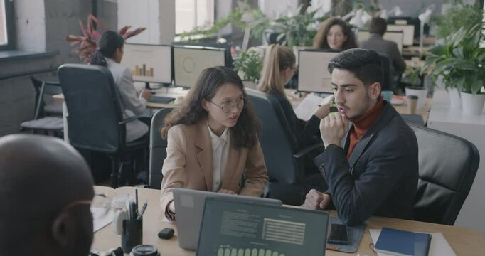Asian woman and Middle Eastern man talking looking at computer screen discussing business while people working at desks. Job and coworking concept.