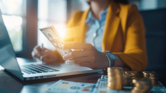 Close-up of a woman in a yellow blazer counting cash with a laptop in front, symbolizing financial management, savings, and investment.