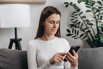 Cute young caucasian woman using smartphone sitting on comfortable sofa at home. Smiling beautiful girl using cell phone and texting. Technology, startup, new business and communication concept