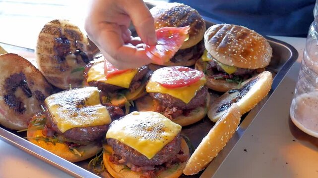 Making burgers and hamburgers outdoor.  Cook preparing cheesebugres. Male puts tomatoes down the cheeseburger