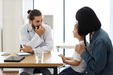 Obraz premium Caucasian male doctor discusses child's health with young girl and mother in clinic. Doctor attentively listens as mother comforts daughter during visit.