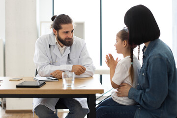 Caucasian male doctor consults young Caucasian girl with mother in medical clinic. Doctor listens attentively as girl discusses health concerns.