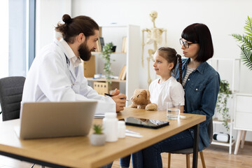 Caucasian male doctor interacts with young girl holding teddy bear during medical consultation in clinic. Mother, Caucasian woman, listens attentively beside daughter.