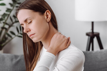 Close up of tired unhappy young woman massaging rubbing stiff sore shoulder tensed muscles fatigued, feeling hurt joint back pain ache sitting alone on sofa at home. Fibromyalgia concept