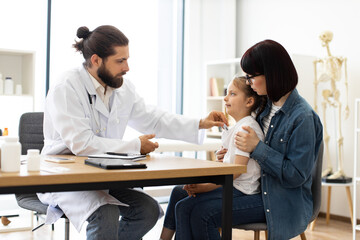 Caucasian doctor measures temperature of little sick girl on woman's lap. Scene set in medical office with professional consultation.