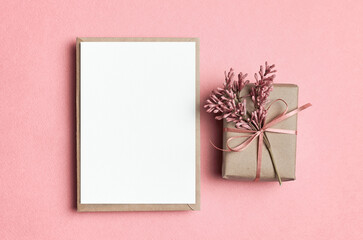 Blank greeting card mockup, white card with envelope, gift box and flowers on pink background