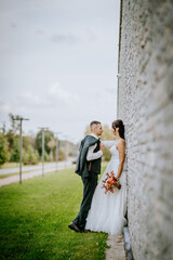 Naklejka premium A groom leans against a stone wall while gazing at his bride, who holds a bouquet. They share an intimate, romantic moment outdoors with greenery around..