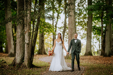 Bride and groom holding hands, walking down a tree-lined path in a serene, green forest setting, both smiling, with the bride holding a bouquet..