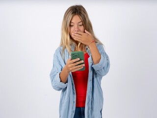 Beautiful teen girl wearing denim jacket being deeply surprised, stares at smartphone display,...