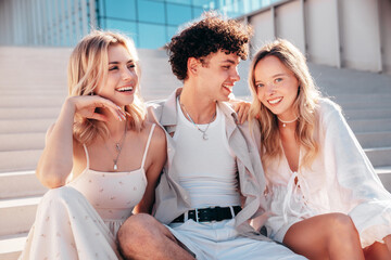 Young three stylish friends posing in the street. Fashion man and two cute female dressed in casual summer clothes. Smiling models having fun. Cheerful women and guy outdoors, in sunny day, at stairs