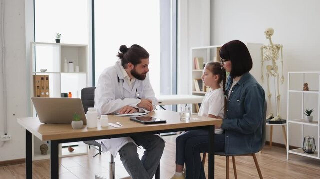 Caucasian doctor examines young girl who is coughing in clinic. Doctor is writing prescription while mother holds girl on lap. Scene depicts healthcare and care in medical setting.