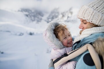 Fototapeta premium Mother standing in the middle of snowy landscape with daughter in baby carrier. Mom and girl enjoying winter holiday in the mountains.