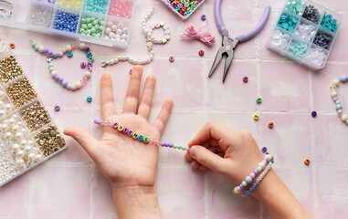 Child making beaded bracelets at home on pink tiled table