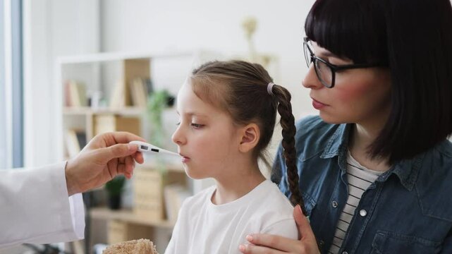 Caucasian doctor measures temperature of young Caucasian girl who appears unwell. Caring mother stands by, providing comfort to her daughter. Scene conveys warmth, care, and concern in medical setting