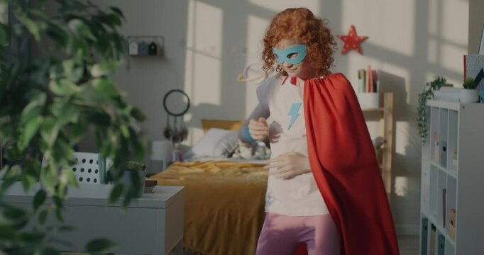 Slow motion of playful little girl in superhero costume mask and cape dancing moving to music having fun at home. Childhood and leisure concept.