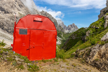 Alpine bivaco mountain hut shelter located in rugged alpine terrain. Red bivacco shelter in Dolomites. Compact structure against stunning landscapes, surrounded by peaks, valleys, and nature.