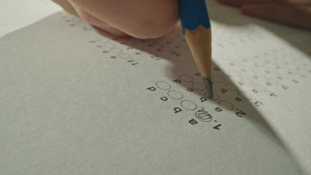 Macro close up of Unrecognizable hands filling up answer sheet with a pencil in classroom. Student shading circles correct answers in standard multiple choice exam test paper. Educational questions.