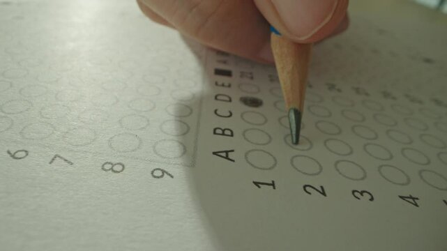 Macro close up of Unrecognizable hands filling up answer sheet with a pencil in classroom. Student shading circles correct answers in standard multiple choice exam test paper. Educational questions.