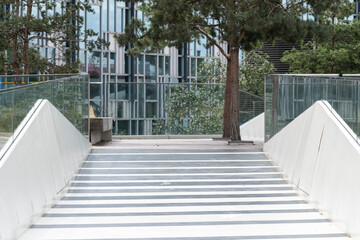 A minimalist pedestrian bridge design incorporating sleek glass railings with trees visible in the background, creating a harmonious blend of urban and natural elements.