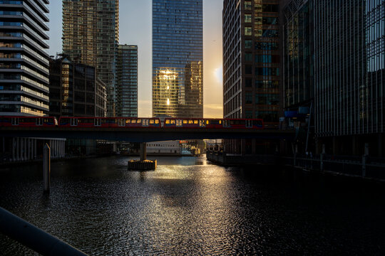 In a stunning contrast of light and shadow, a vibrant red train crosses a bridge over a tranquil waterway, backlit by a radiant golden sunset in the city in London UK