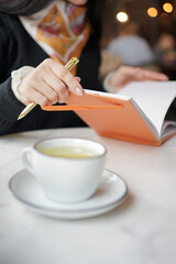The girl makes notes in a notebook while sitting at the table with a cup of tea.