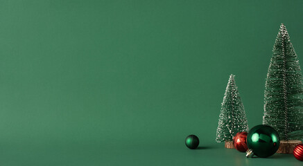 A minimalist setup featuring small Christmas trees and colorful baubles against a green backdrop, evoking holiday spirit