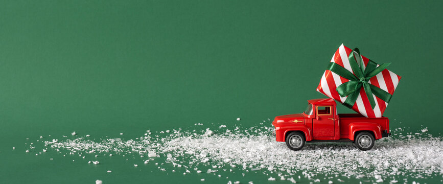 A red toy truck carrying a striped gift box with a green bow moves through snow against a green background, evoking festive holiday cheer and a sense of playful joy