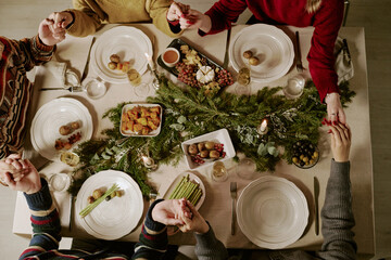 Top down shot of unrecognizable family holding hands while praying, they sitting at table with traditional festive dinner and seasonal decor
