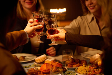 Medium close up of unrecognizable friends clinking glasses with red wine while sitting at table in dimly lit room