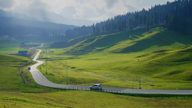 A wide scenic shot of a road passing through a lush green meadow with sunlight casting golden patches on the slopes of the hills and in the background mountains surrounded by the rain clouds