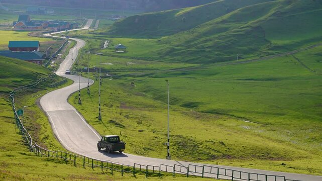 A wide scenic cinematic shot of a pick up van or vehicle driving along a picturesque winding mountain road passing through a lush green meadow with sunlight casting golden patches over a landscape. 