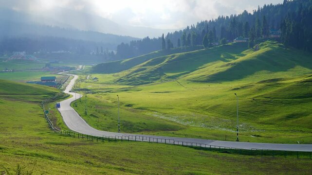 A wide scenic cinematic shot of a picturesque winding mountainous road passing through a lush green meadow with sunlight casting golden patches over landscape with mountains visible in the background