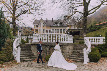Obraz premium A bride and groom are walking down a set of stairs in front of a yellow house. The bride is wearing a white gown and the groom is wearing a suit. The scene is romantic and intimate