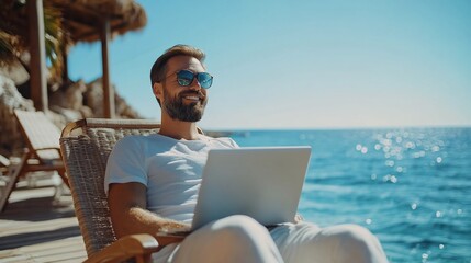 A man is smiling while sitting on a beach chair and working on his laptop.