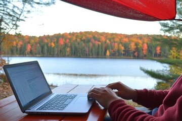Person typing on laptop at outdoor table with lake and autumn trees in background