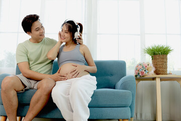 Cheerful Asian couple sit on sofa couch at living room together. Beautiful pregnant wife with headphones enjoys listening to music for relaxation while husband take care of her. Happy family at home.