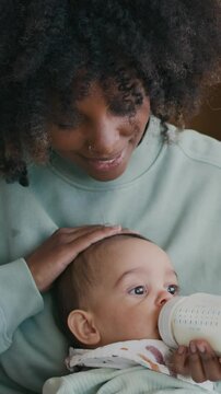 Vertical shot of young African American single mother cuddling her baby on head and feeding him with milk from plastic bottle while holding infant on her knees