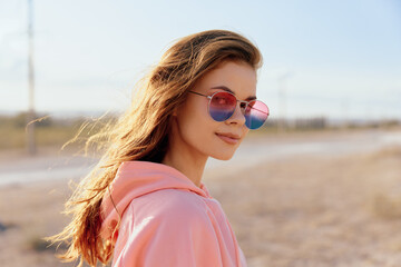 Stylish young woman in pink hoodie and sunglasses standing on desert road under bright blue sky