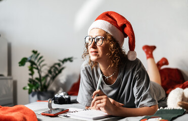 Woman in red cap writing down Christmas goals and plans on the sofa in notebook in cozy home...
